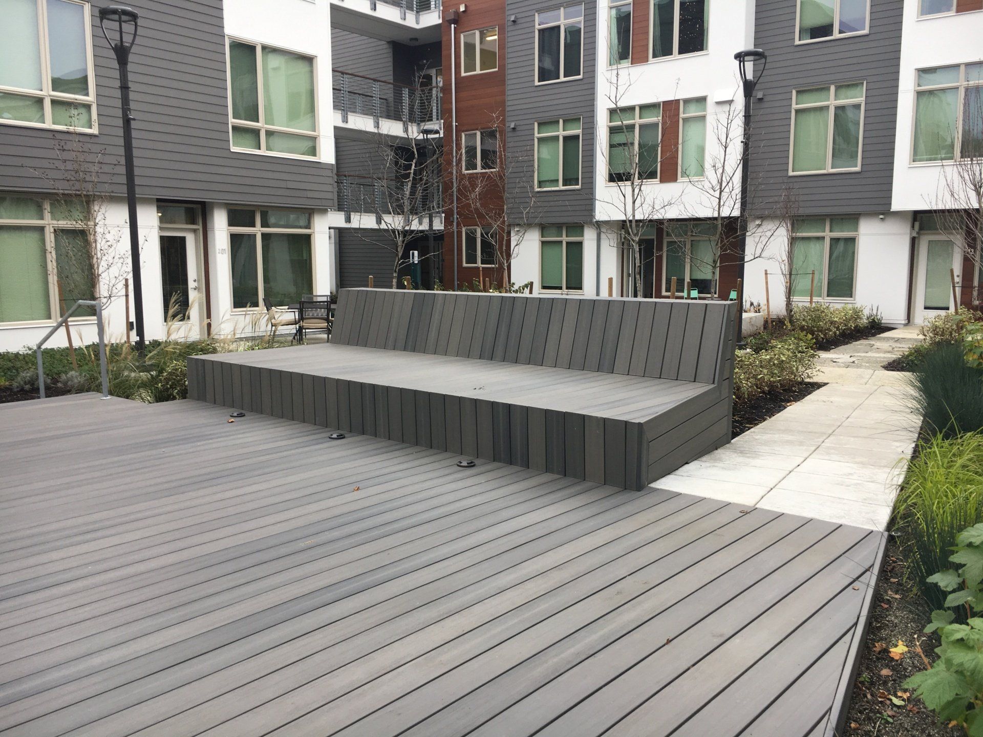 Outdoor seating area with gray wooden deck and bench in front of multi-story building.