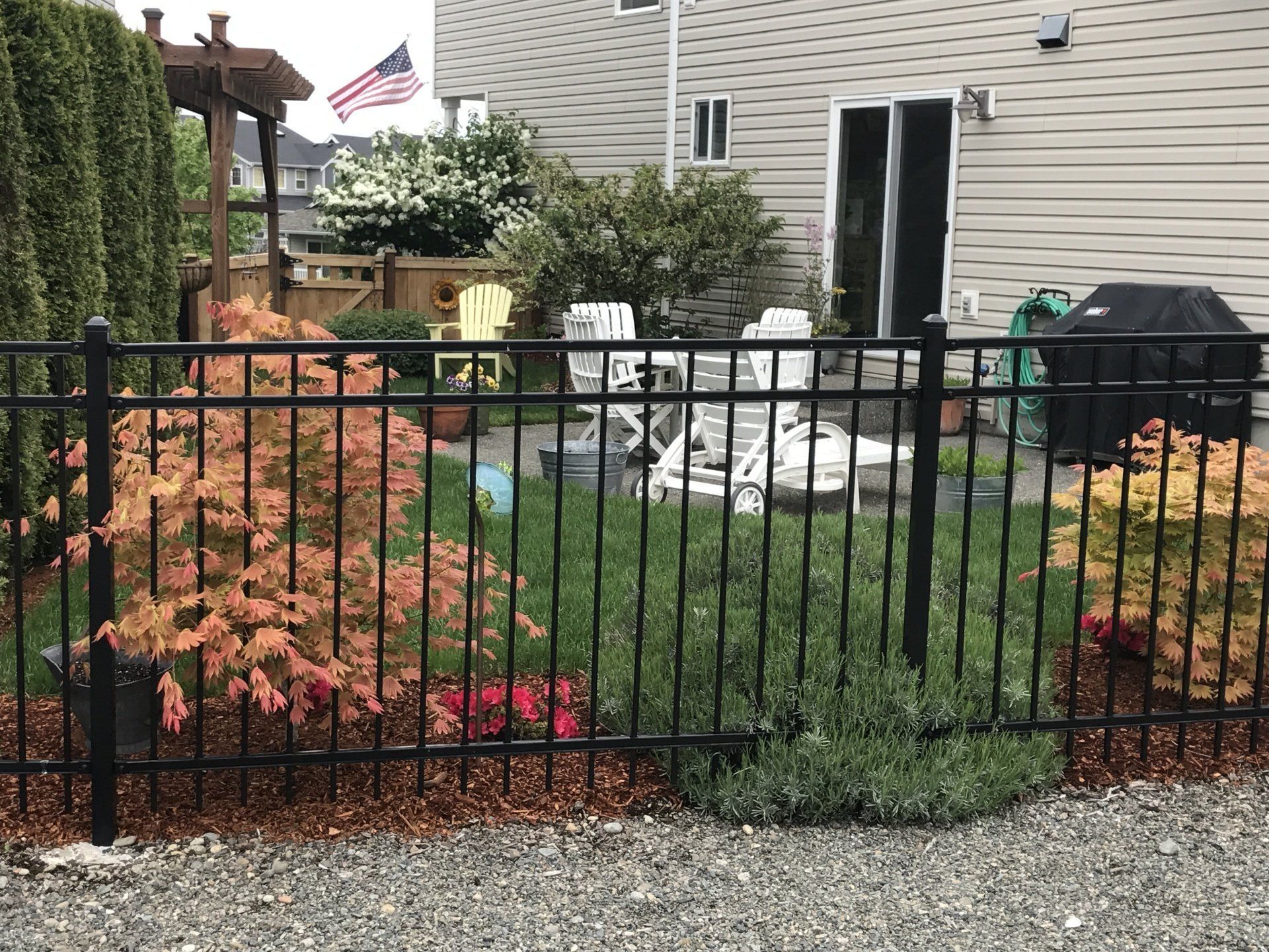 Black wrought-iron fence encloses a backyard with patio, plants, and house. American flag flies.