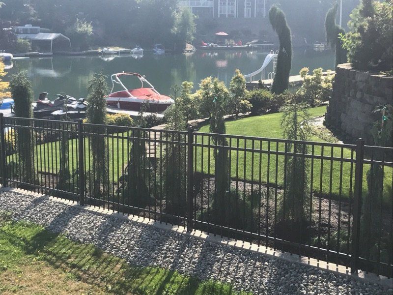 Black metal fence along a green lawn with trees, lake, and boats in the background.