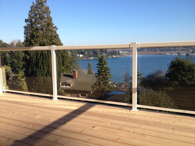 Deck overlooking a lake with glass and white railing. Trees and houses visible. Sunny day.