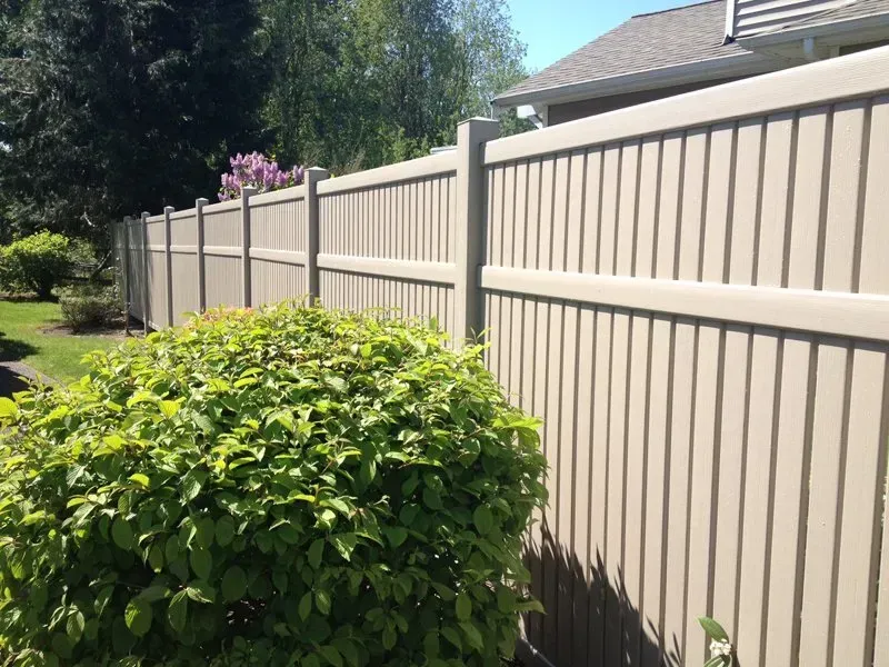 Beige vinyl privacy fence in a yard with green shrubbery and trees.