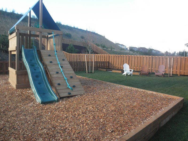 Playground with two slides, climbing wall, and wood chip base, next to a grassy area.