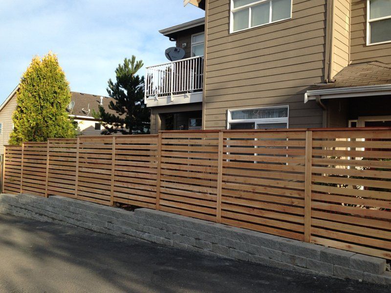 Wooden horizontal slat fence in front of a tan two-story building on a sunny day.