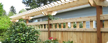 Wooden pergola over a fence in a backyard setting. Green foliage and a blue house are visible.