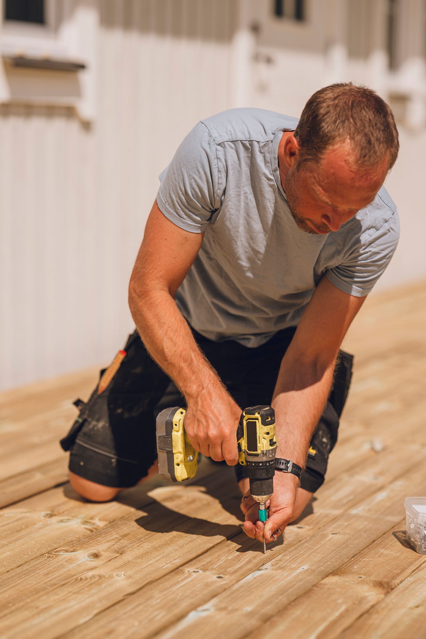 Man kneeling on wooden deck, using a power drill. Outdoors, sunny.