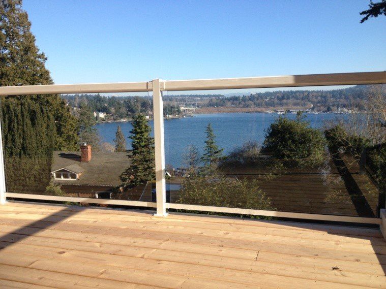 Wooden deck with glass railing overlooking a blue lake and trees under a clear sky.