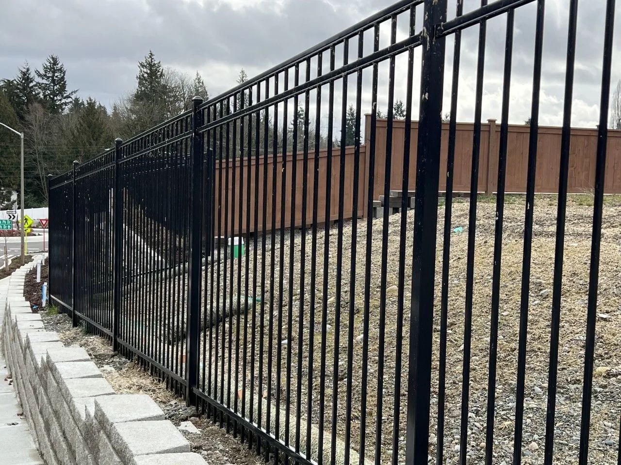 Black metal fence along a gray retaining wall, with a brown wall in the background. Cloudy sky.