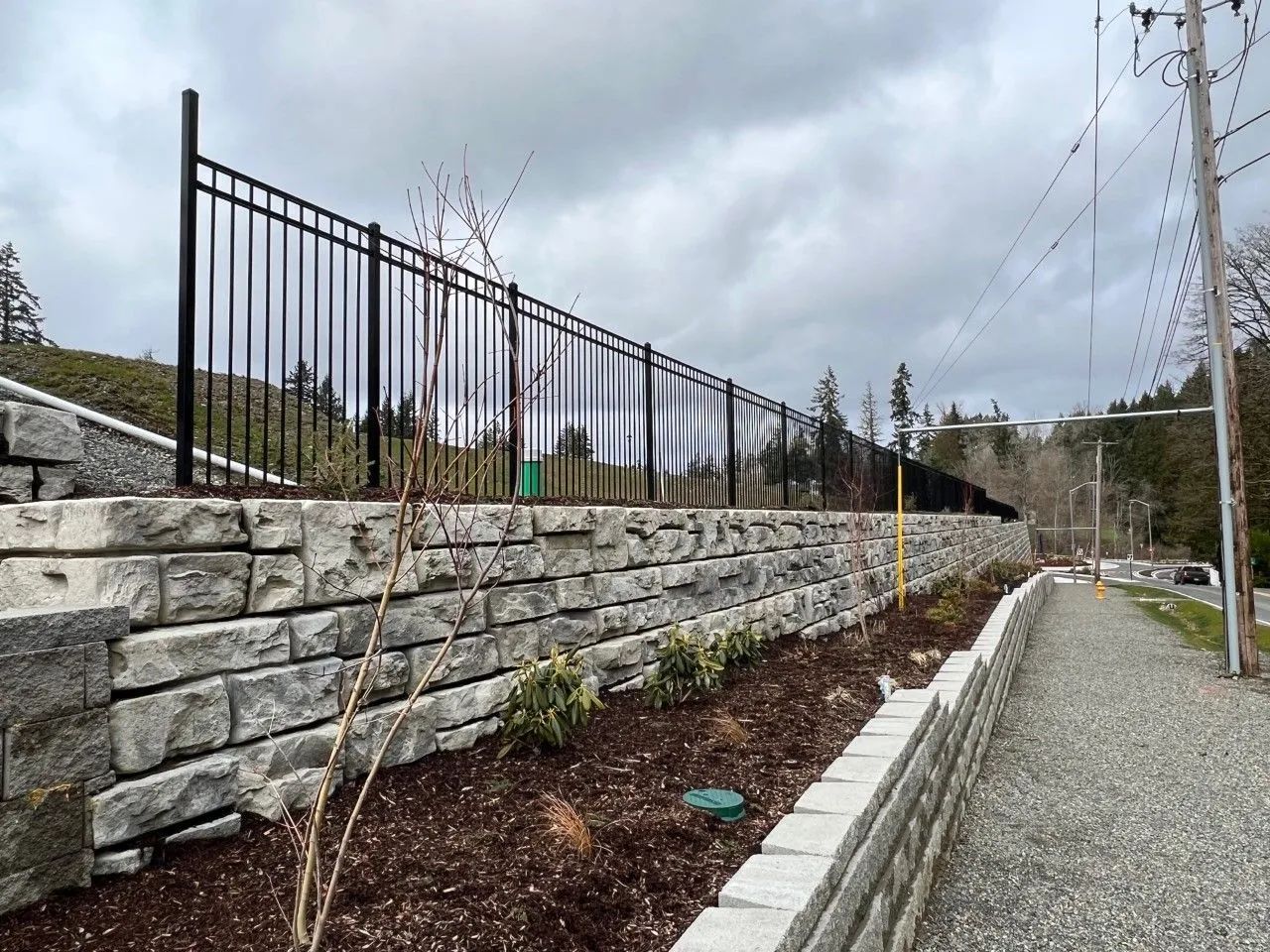 Black metal fence atop a stacked stone retaining wall. Gravel path beside the wall. Overcast sky.