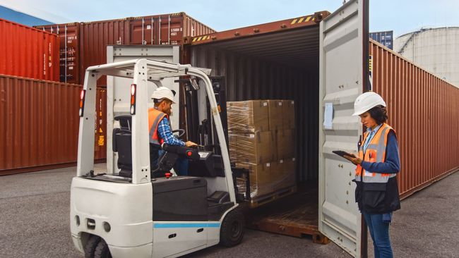 A man is driving a forklift in a warehouse while a woman looks on.