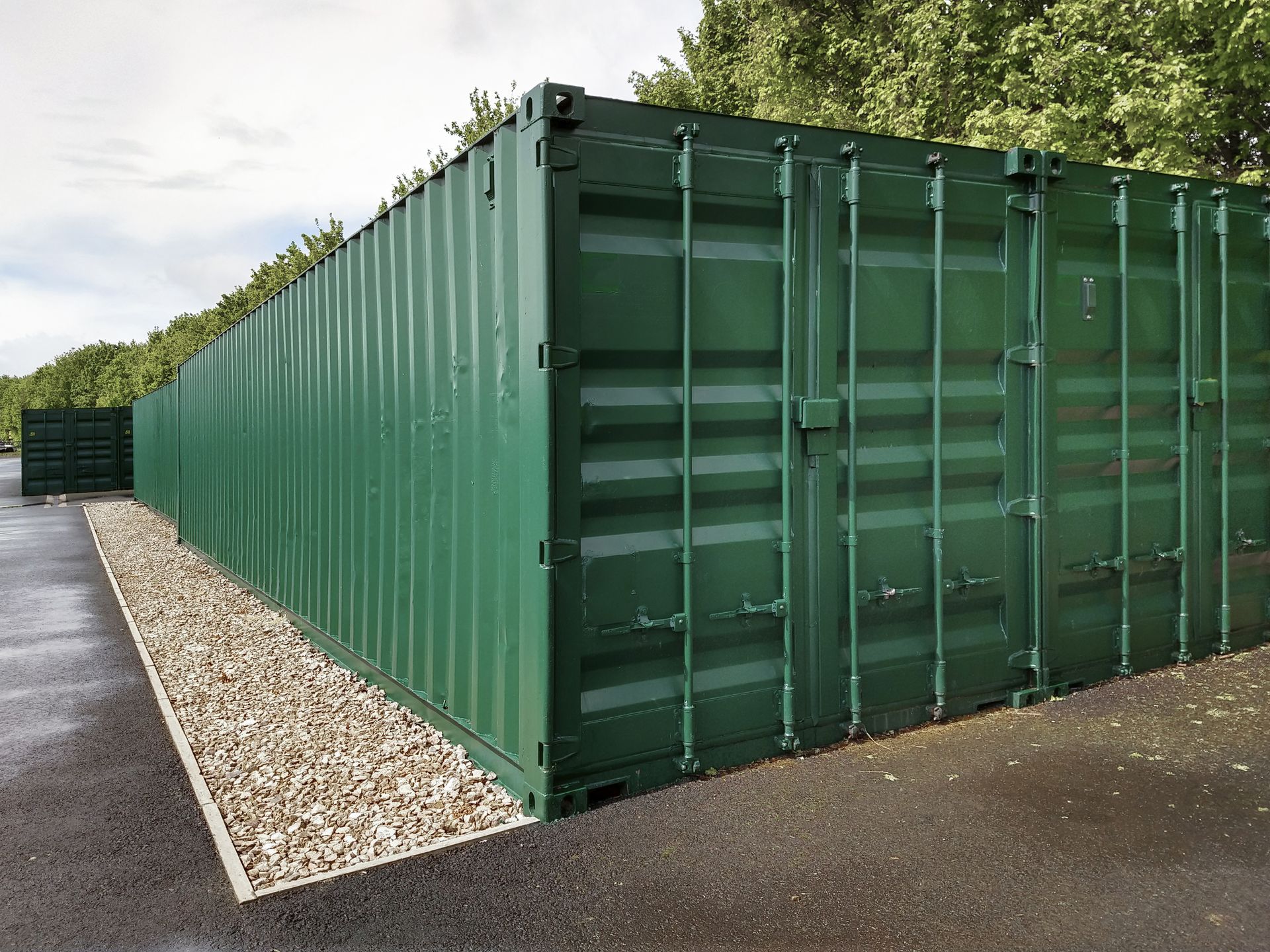 A row of green shipping containers are lined up on the side of a road.