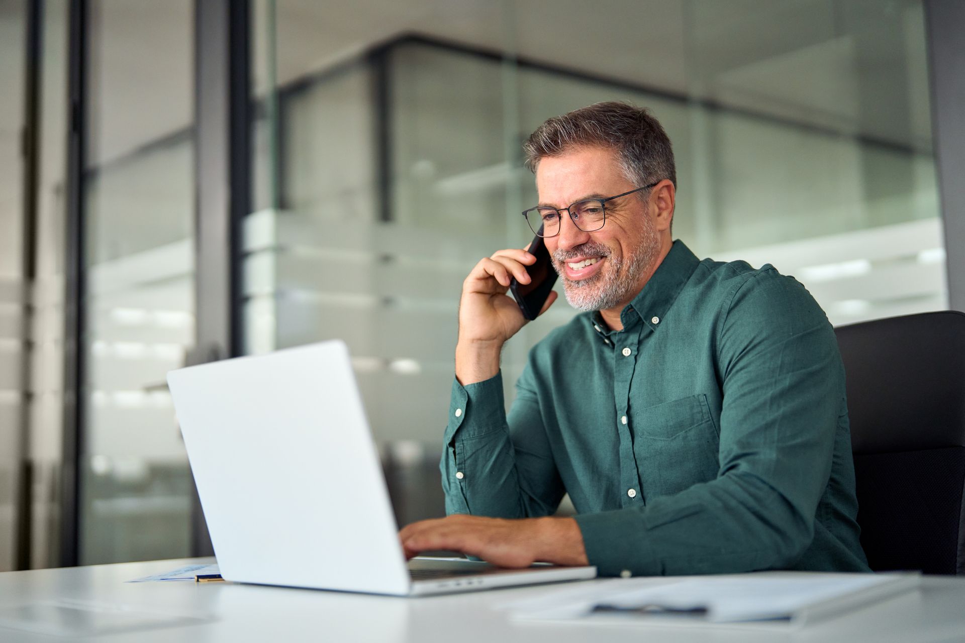 A man is talking on a cell phone while using a laptop computer.