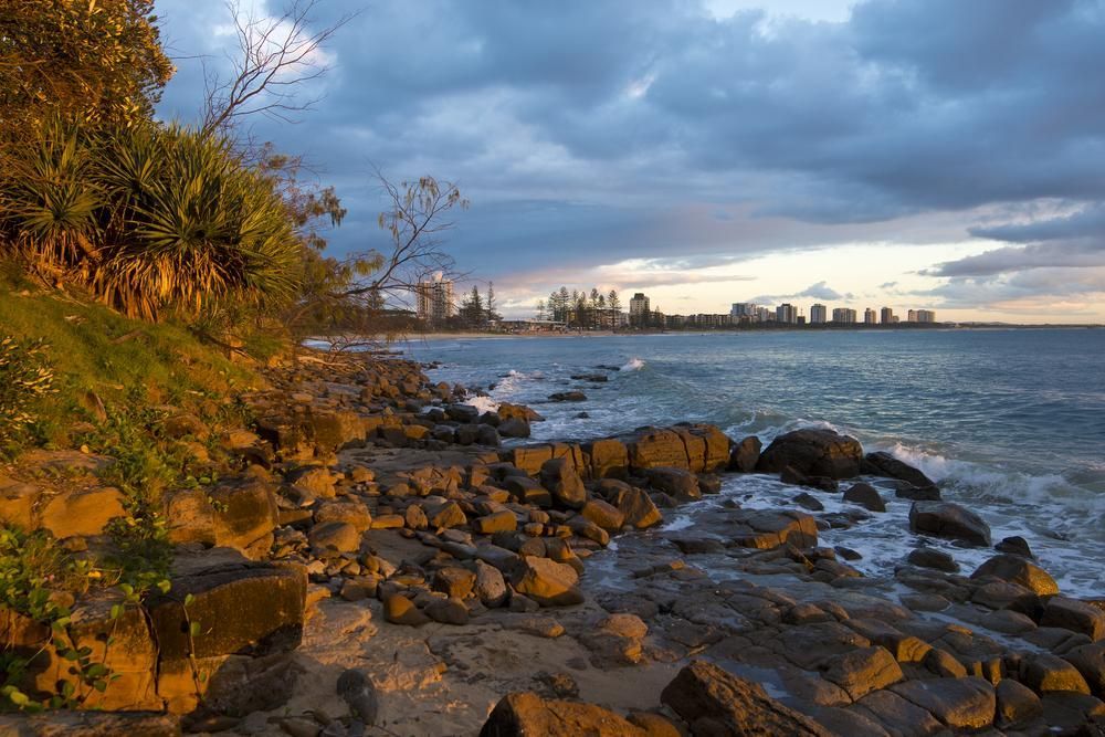 A Rocky Beach With a City in the Background at Sunset — Abbsolve Services in Maroochydore, QLD