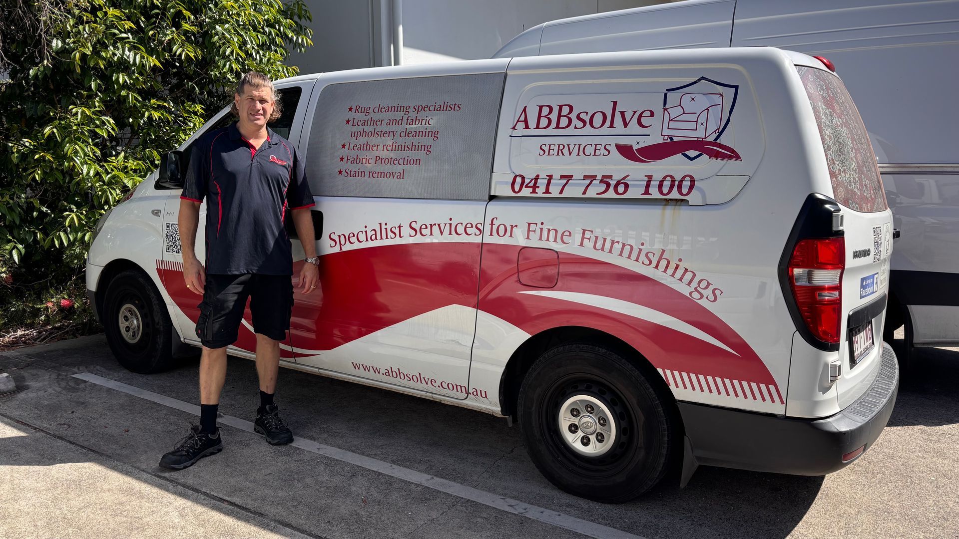 Man carrying a large rolled carpet in a workshop, wearing dark clothes and boots.