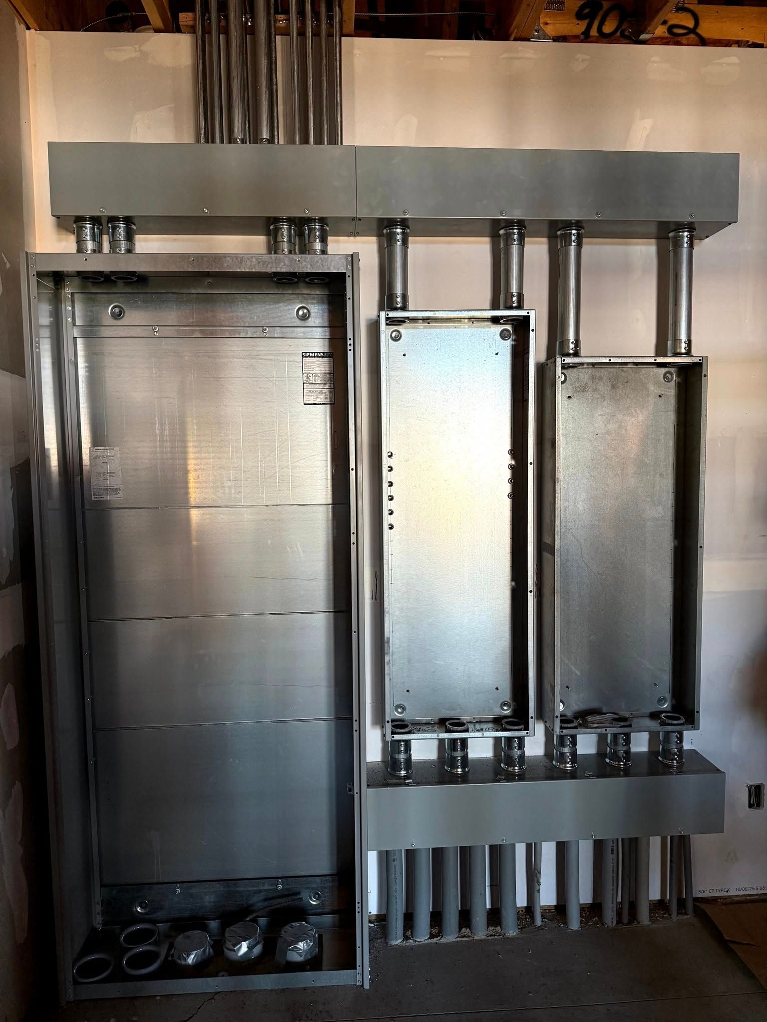 Electrical panel with three connected sections, conduits above and below, against a wall.