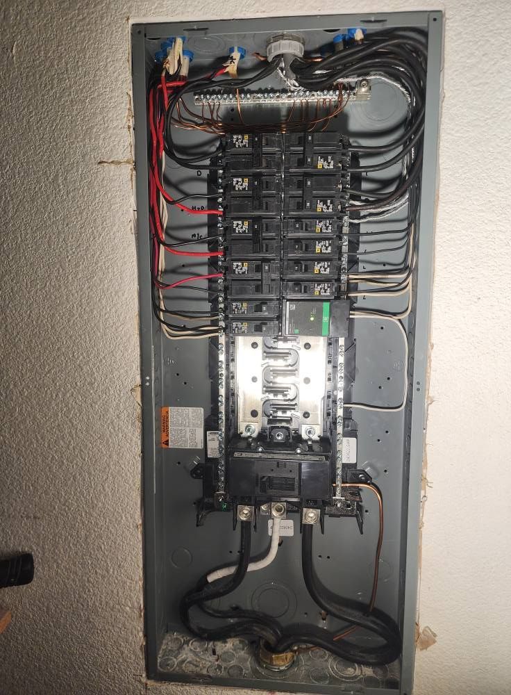 Electrical panel with numerous black wires, gray metal box, and circuit breakers against a white wall.