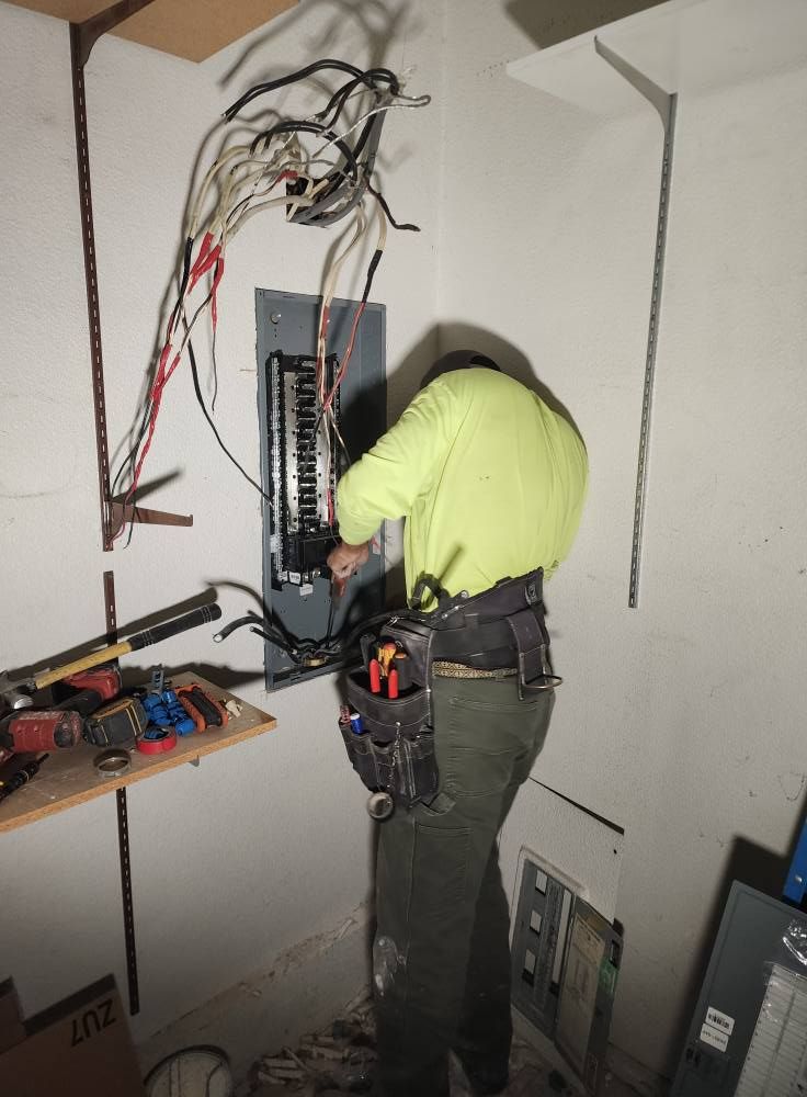 Electrician in lime green shirt working on electrical panel in a small room with tools and wiring.