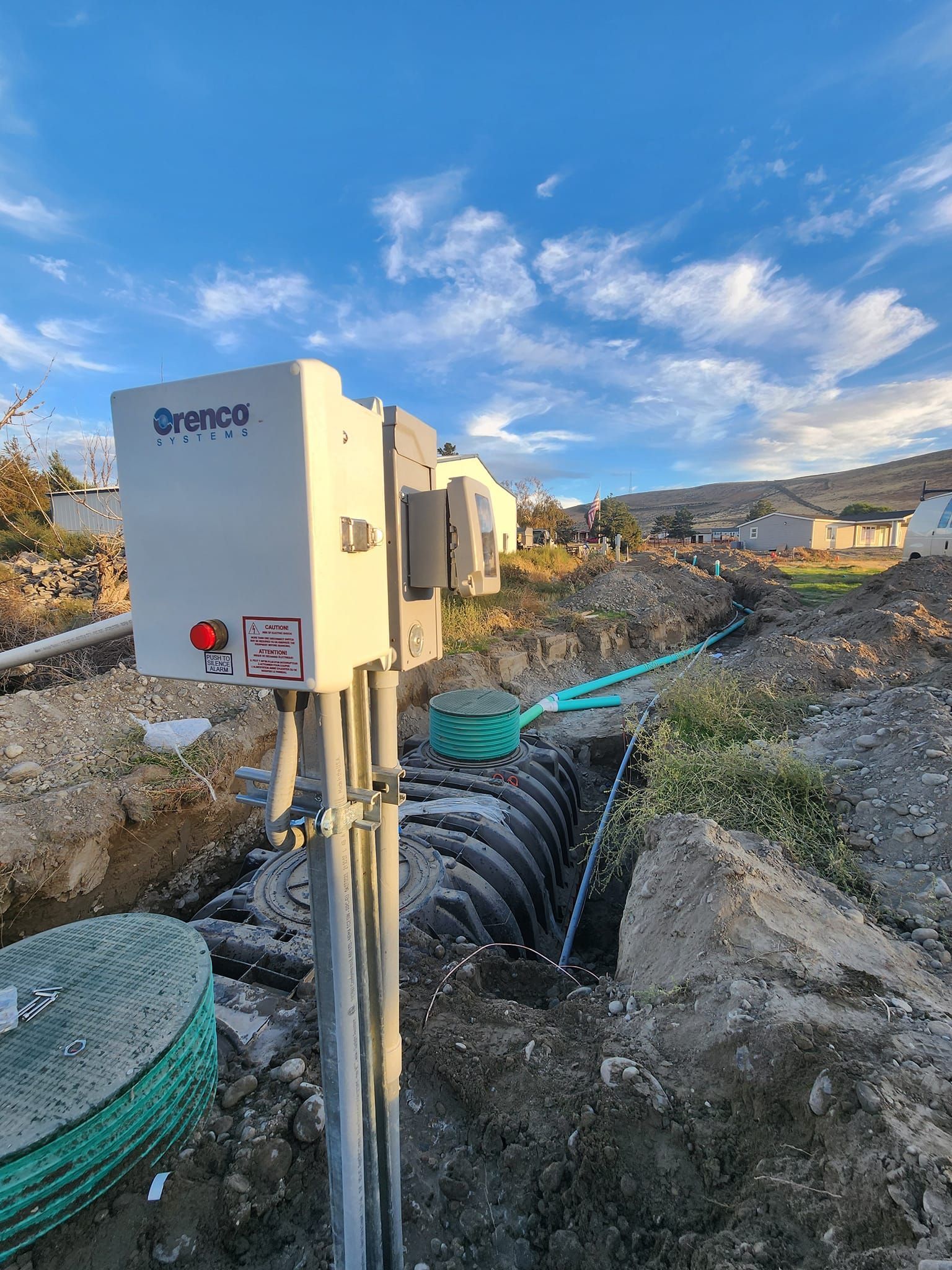 Septic system installation; gray electrical box, black pipe, and green tubing in a dirt trench, under a blue sky.
