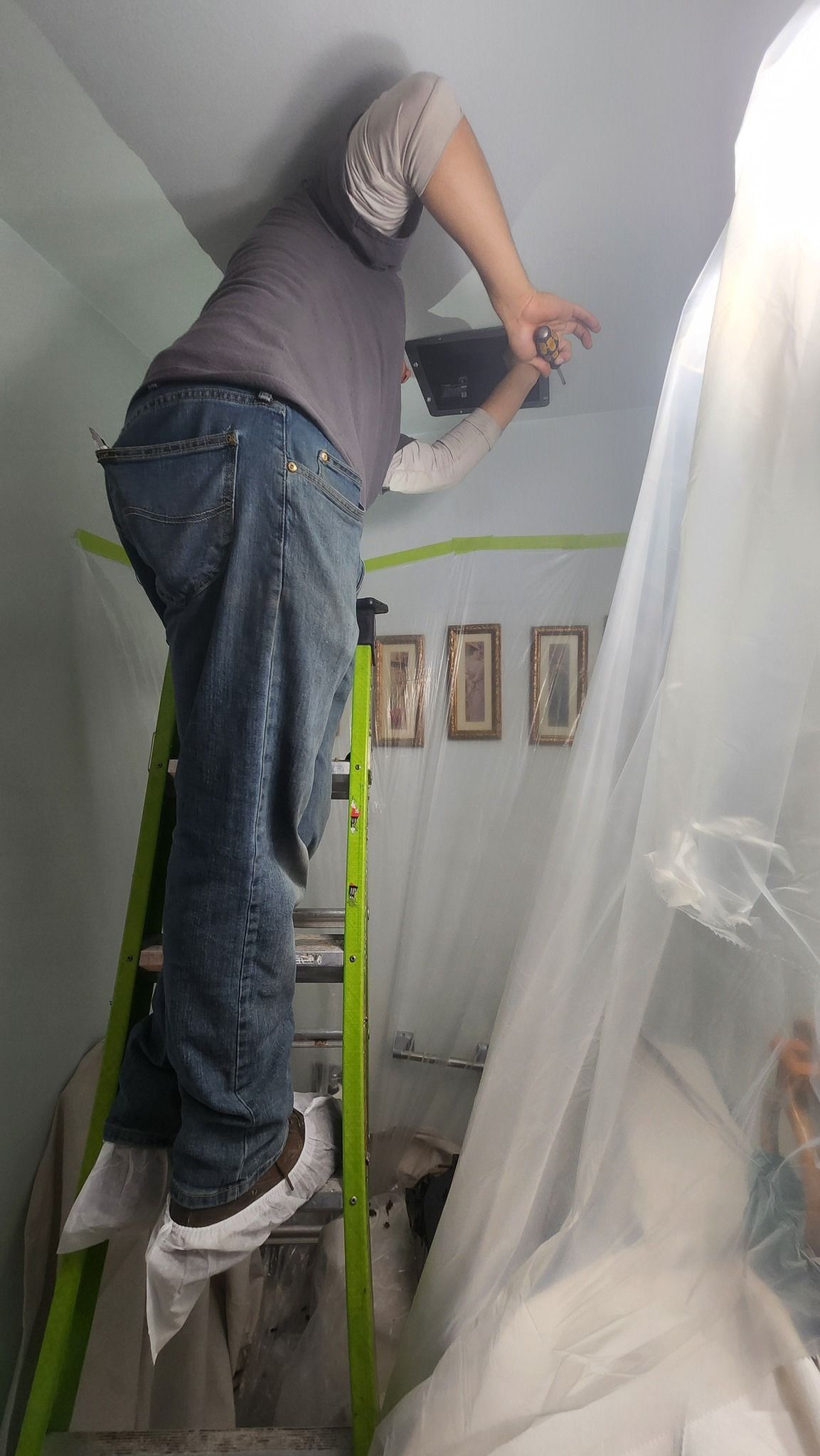 Person on ladder near ceiling, installing something. White protective shoe covers, green ladder, white net on the right.