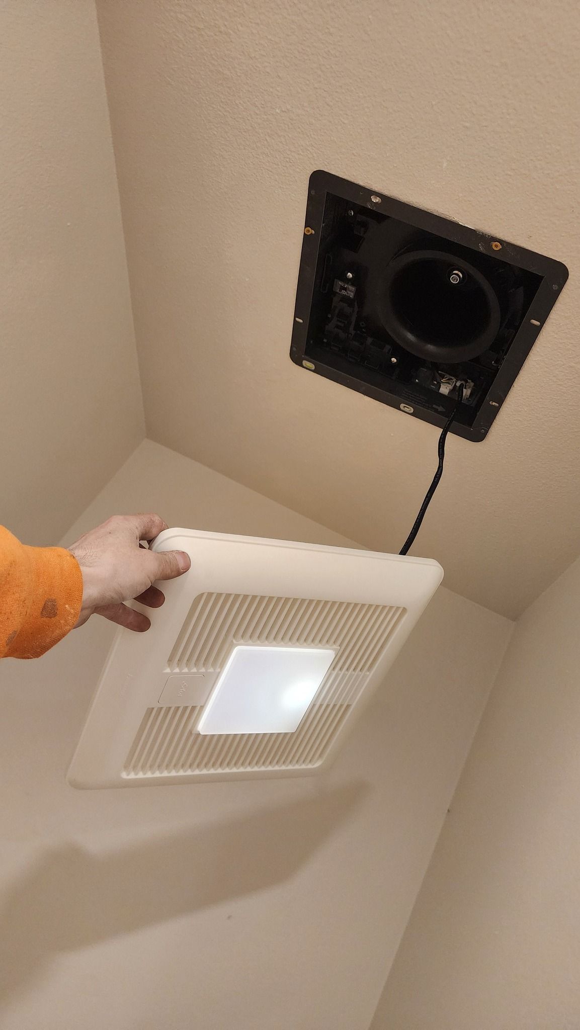 Person holding a white bathroom fan cover near the opened ceiling fixture.