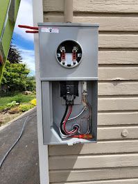 Gray electrical meter box mounted on beige siding; exposed wiring.