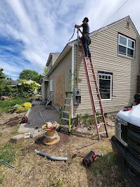 Person on a ladder, installing electrical wiring on a house with tan siding. Tools and wire spool on the ground.