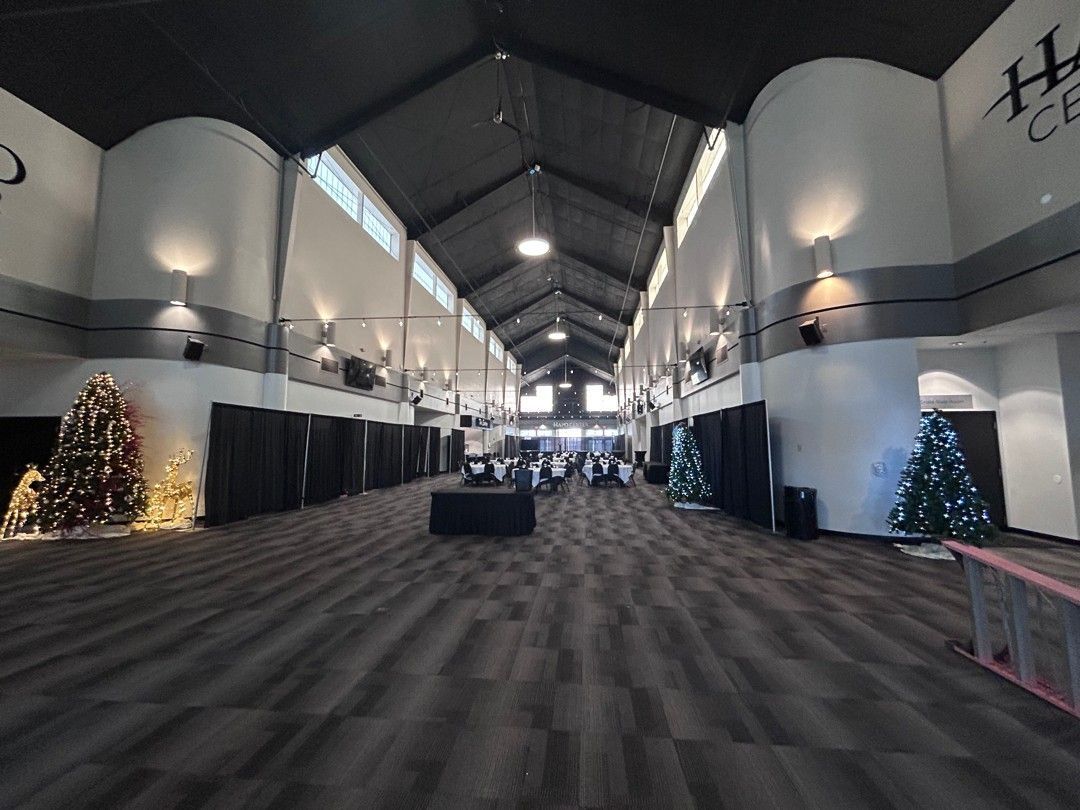 Interior view of a long event space with Christmas trees and tables. Dark gray carpet, white walls.