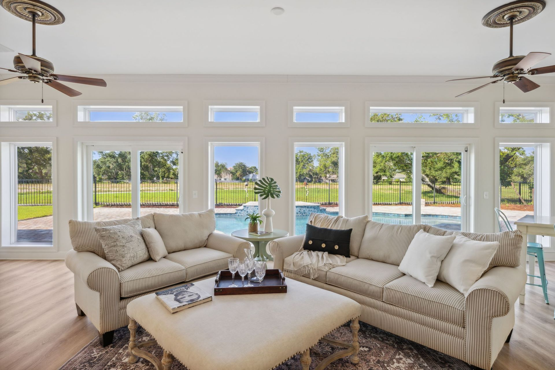 Living room with beige couches, ottoman, large windows overlooking a pool and yard, two ceiling fans.