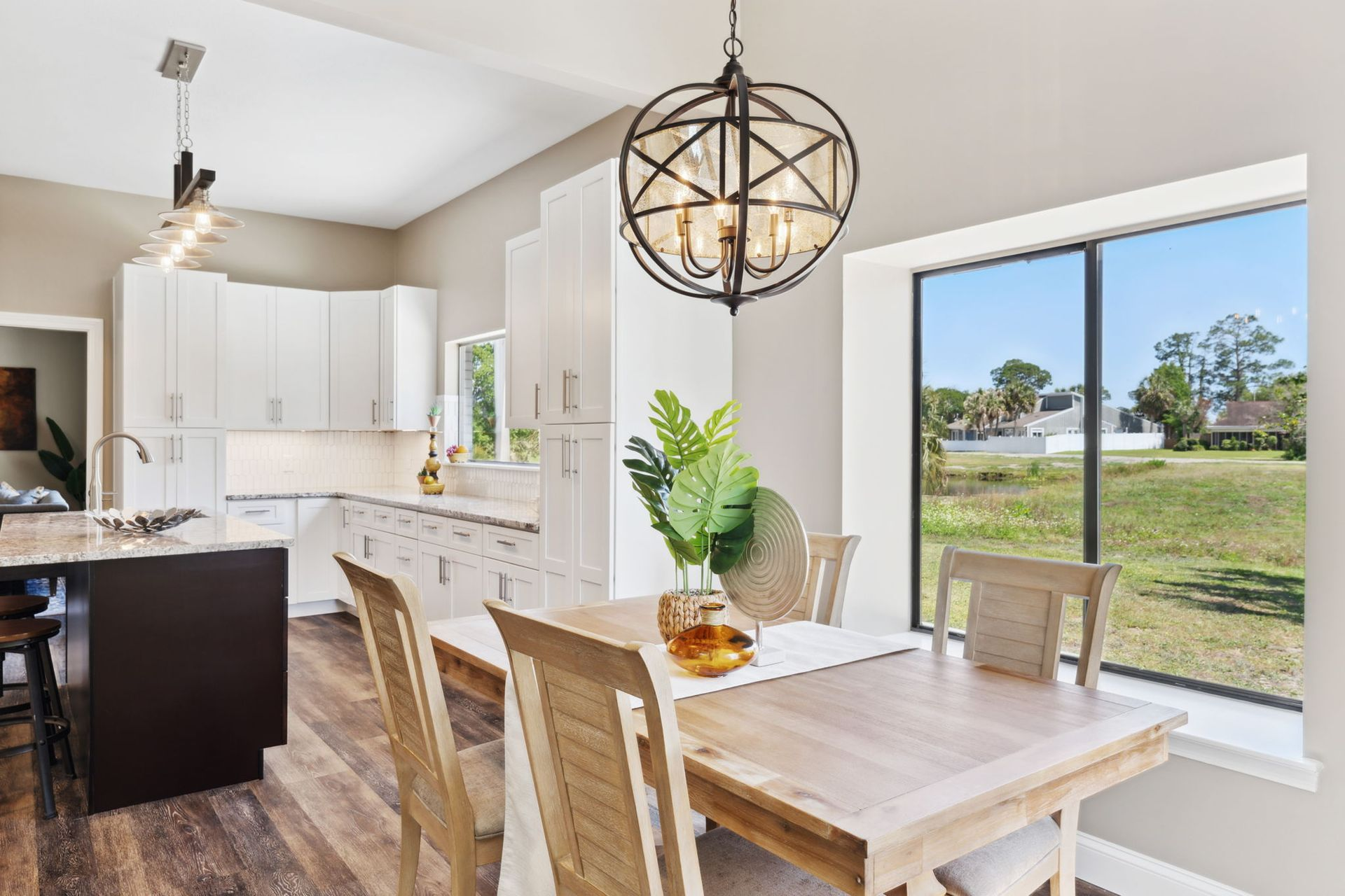 Kitchen and dining area with light wood table, chairs, cabinets, and a dark island. A large window overlooks a grassy yard.