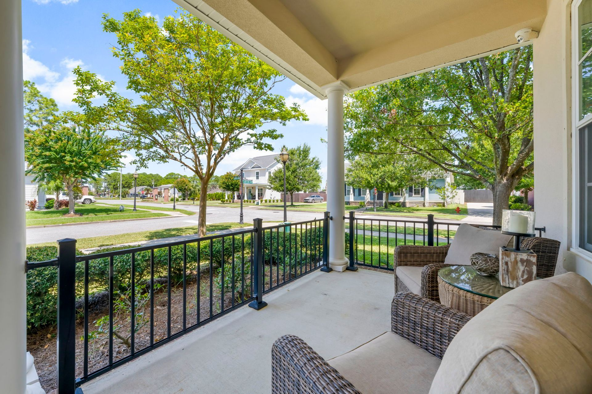 Covered porch with seating overlooking a residential street. Green trees, black railing, and blue sky.