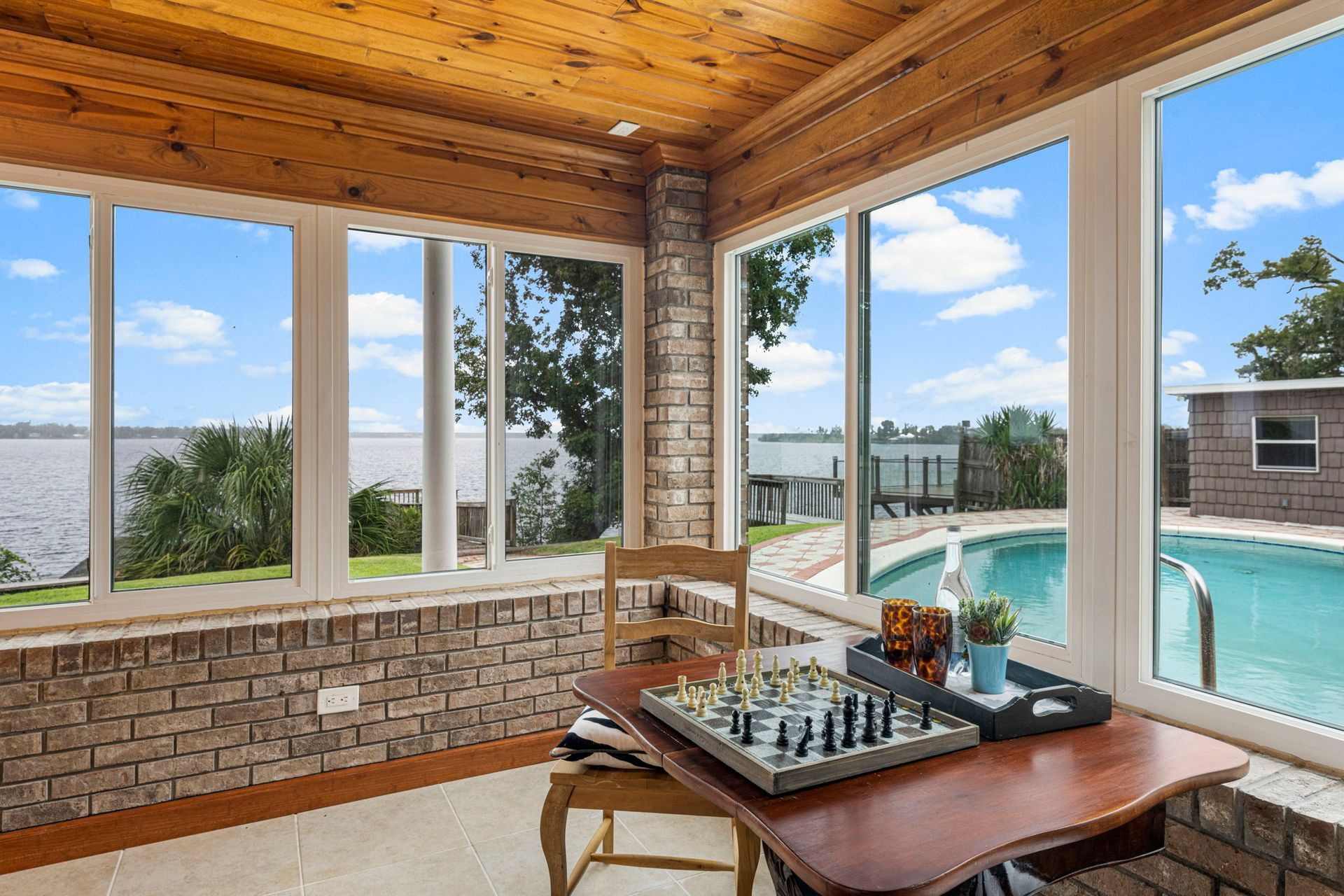 Sunroom with brick walls, wood ceiling, large windows overlooking water and a pool. Chess set on a small table.