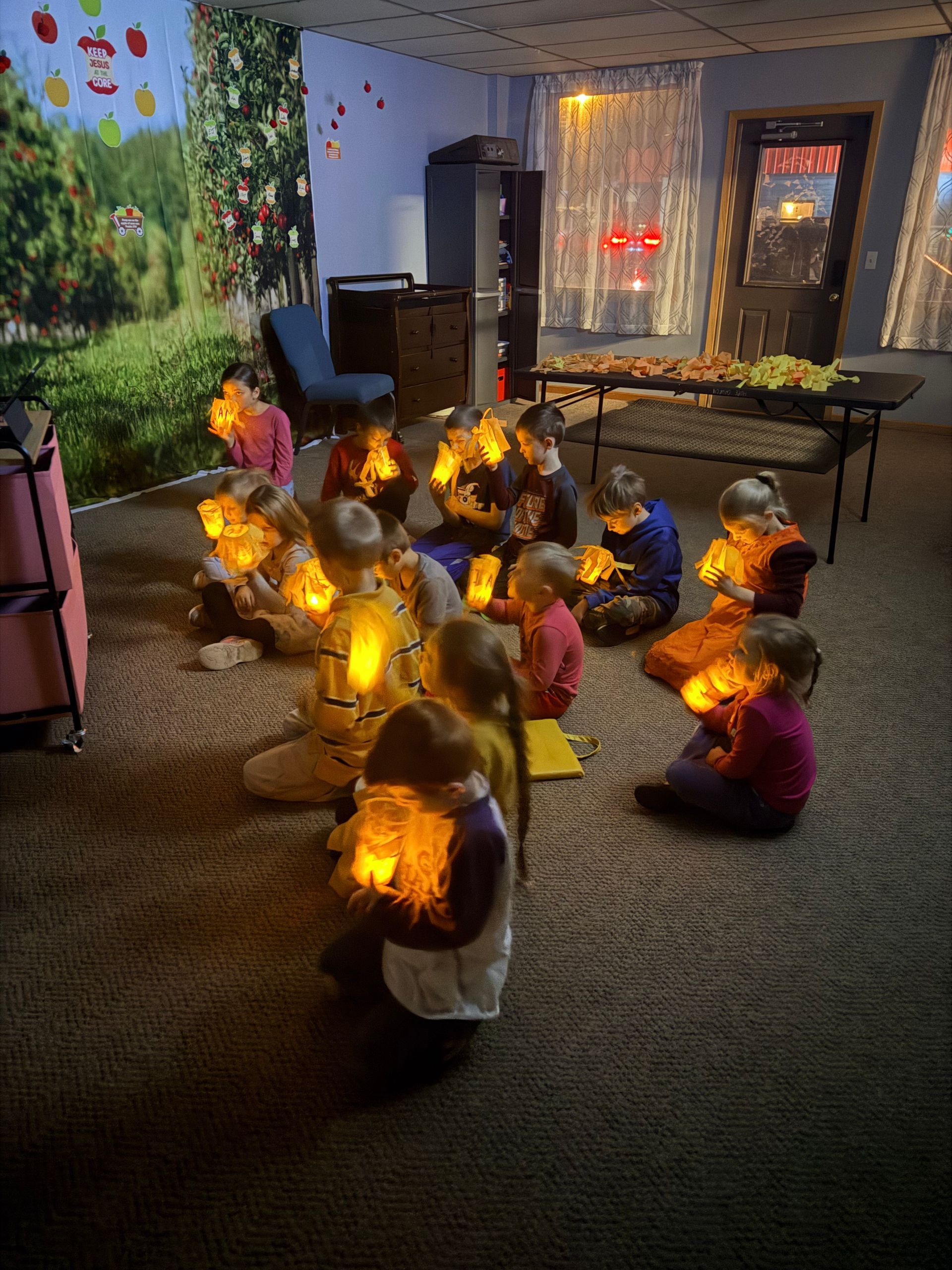 Children seated, holding lit lanterns in a dimly lit room. Background includes a wall mural and a doorway.