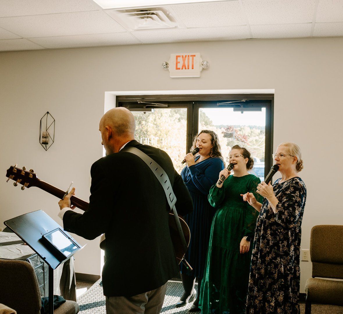 A man playing guitar and three individuals singing into microphones in a room with an exit sign overhead.