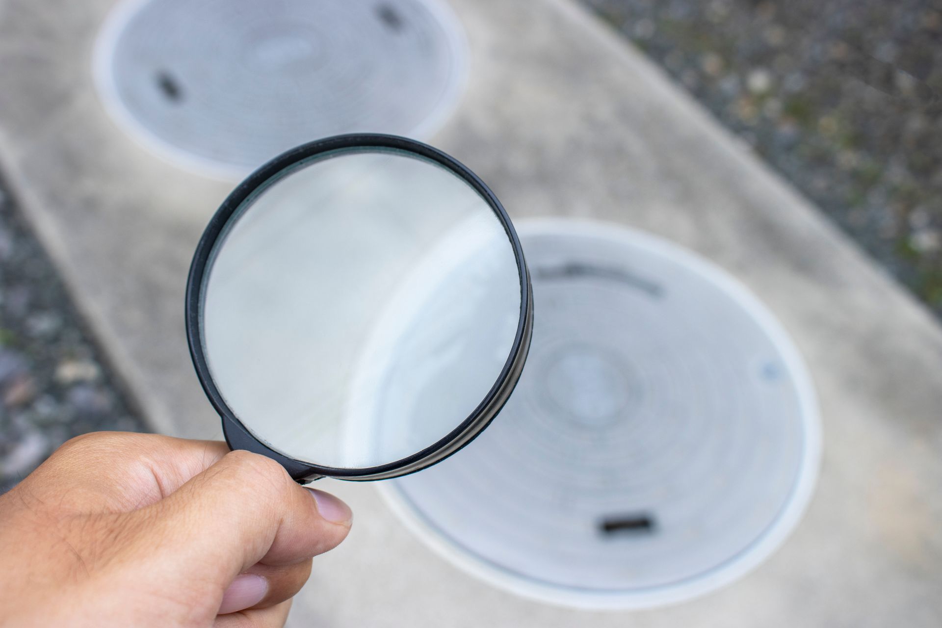 Hand holding magnifying glass over utility access covers on pavement.