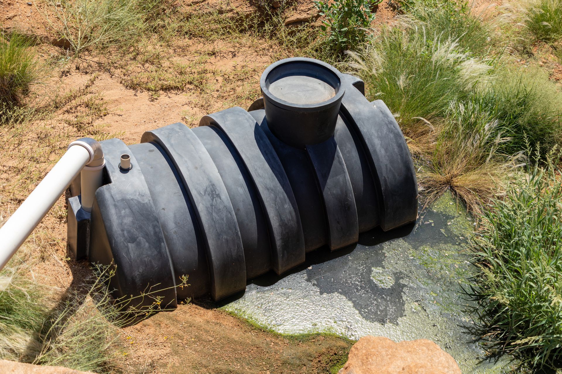 Black, cylindrical water tank in a grassy outdoor setting with connected white pipe.