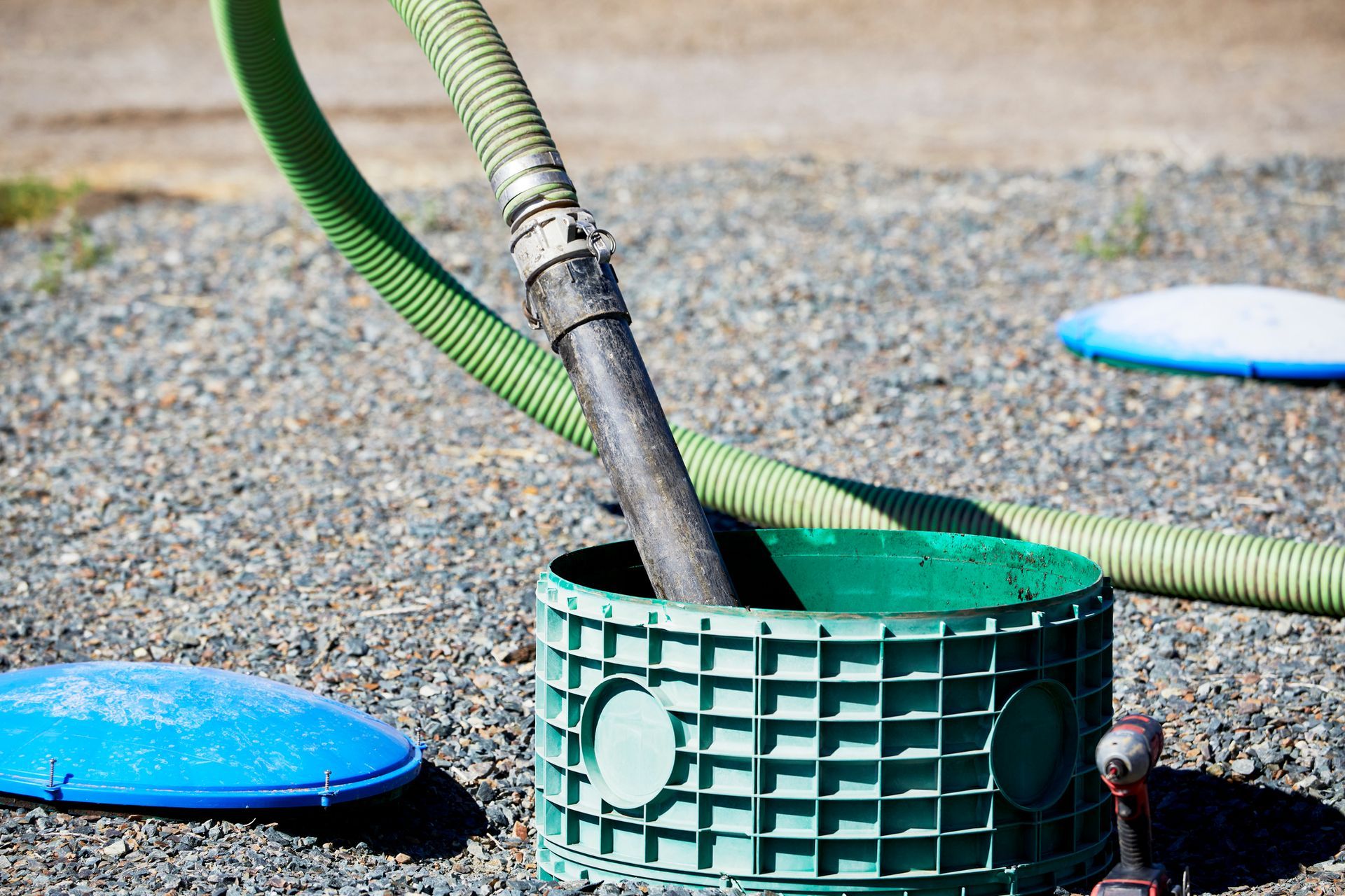 Green septic tank being serviced with hoses on gravel.