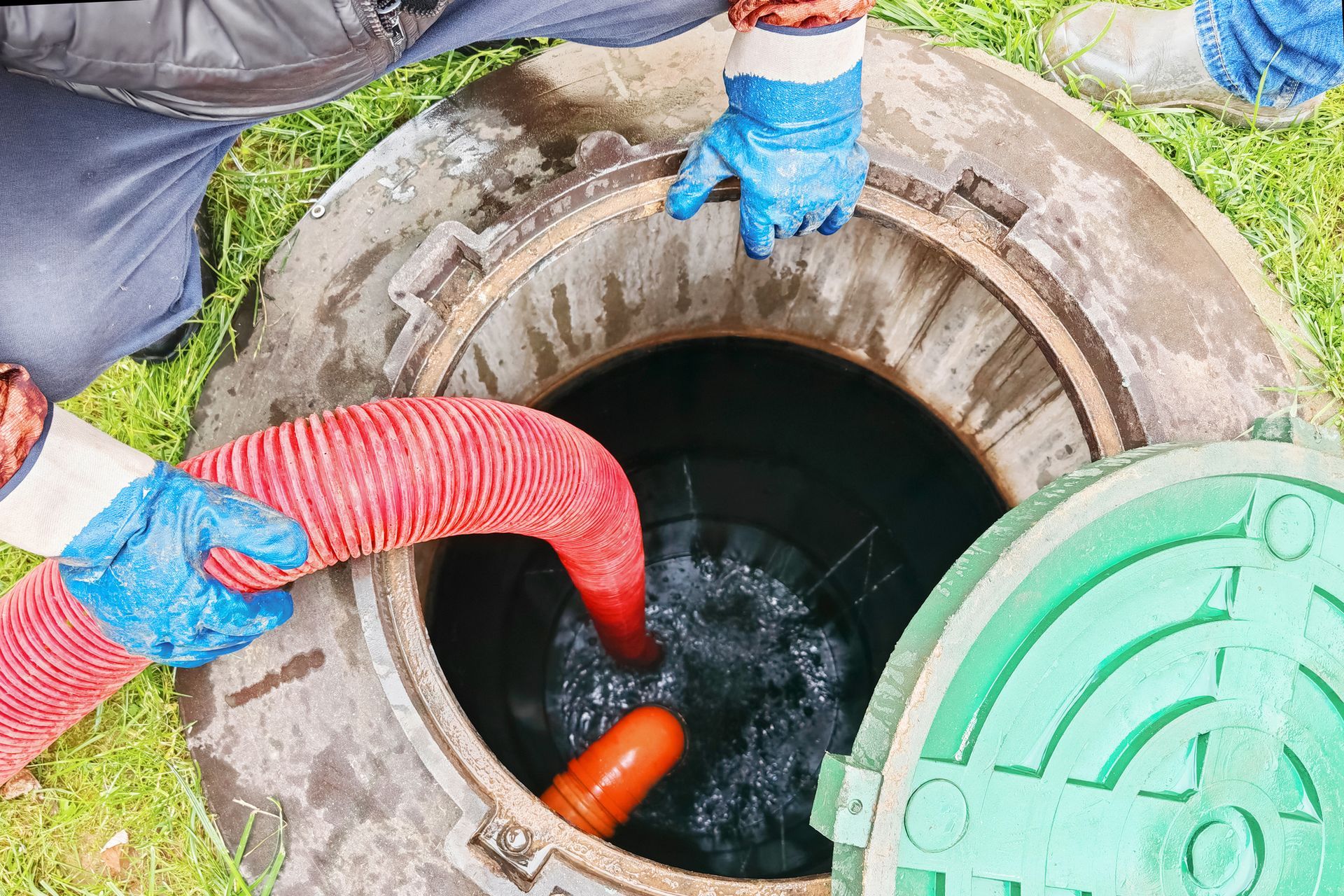 Person pumping a septic tank. Red hose, blue gloves, open concrete manhole, green lid.