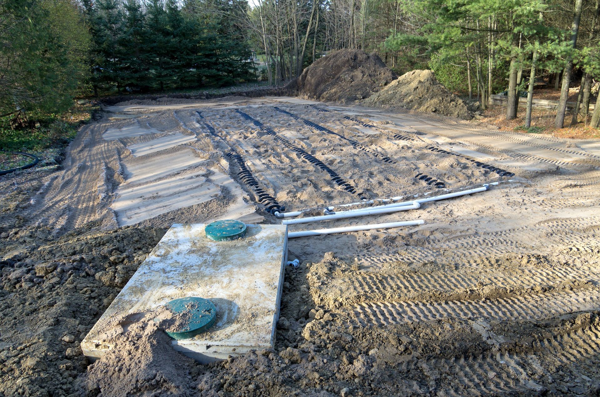 Septic system installation; concrete tank with two lids, soil trenches, pipes, and trees in the background.