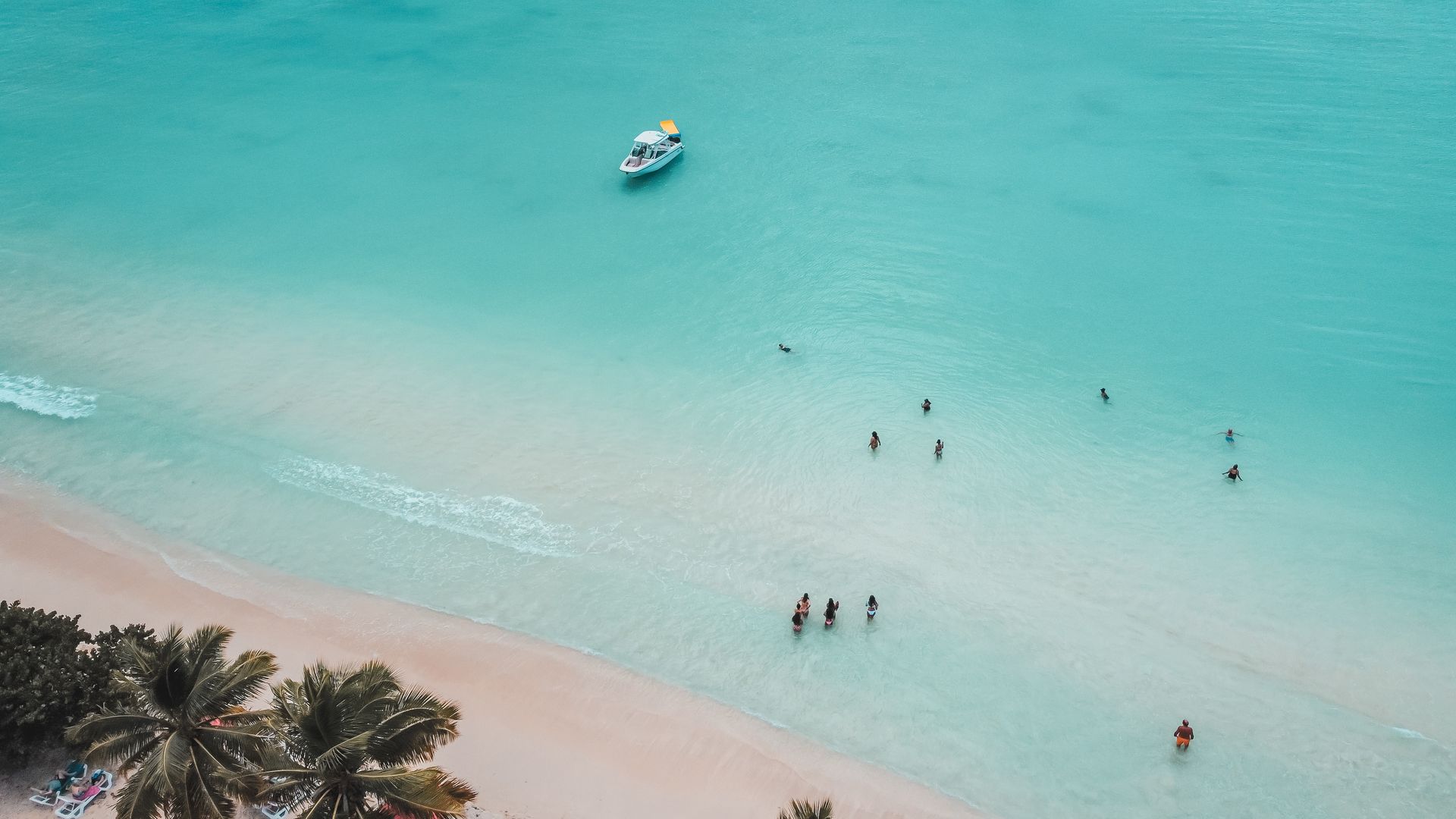 Aerial view of turquoise water, a sandy beach with people swimming and a boat; palm trees in the foreground.