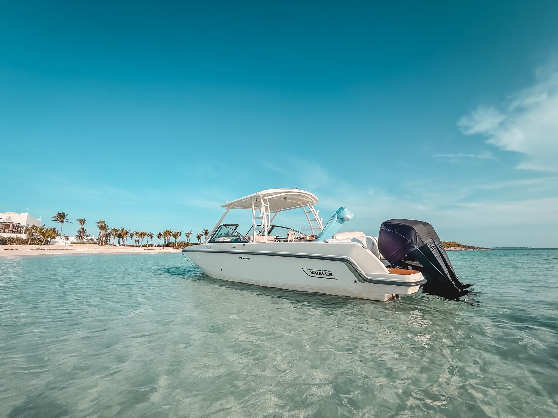 White boat in clear, turquoise water near a beach with palm trees under a bright blue sky.
