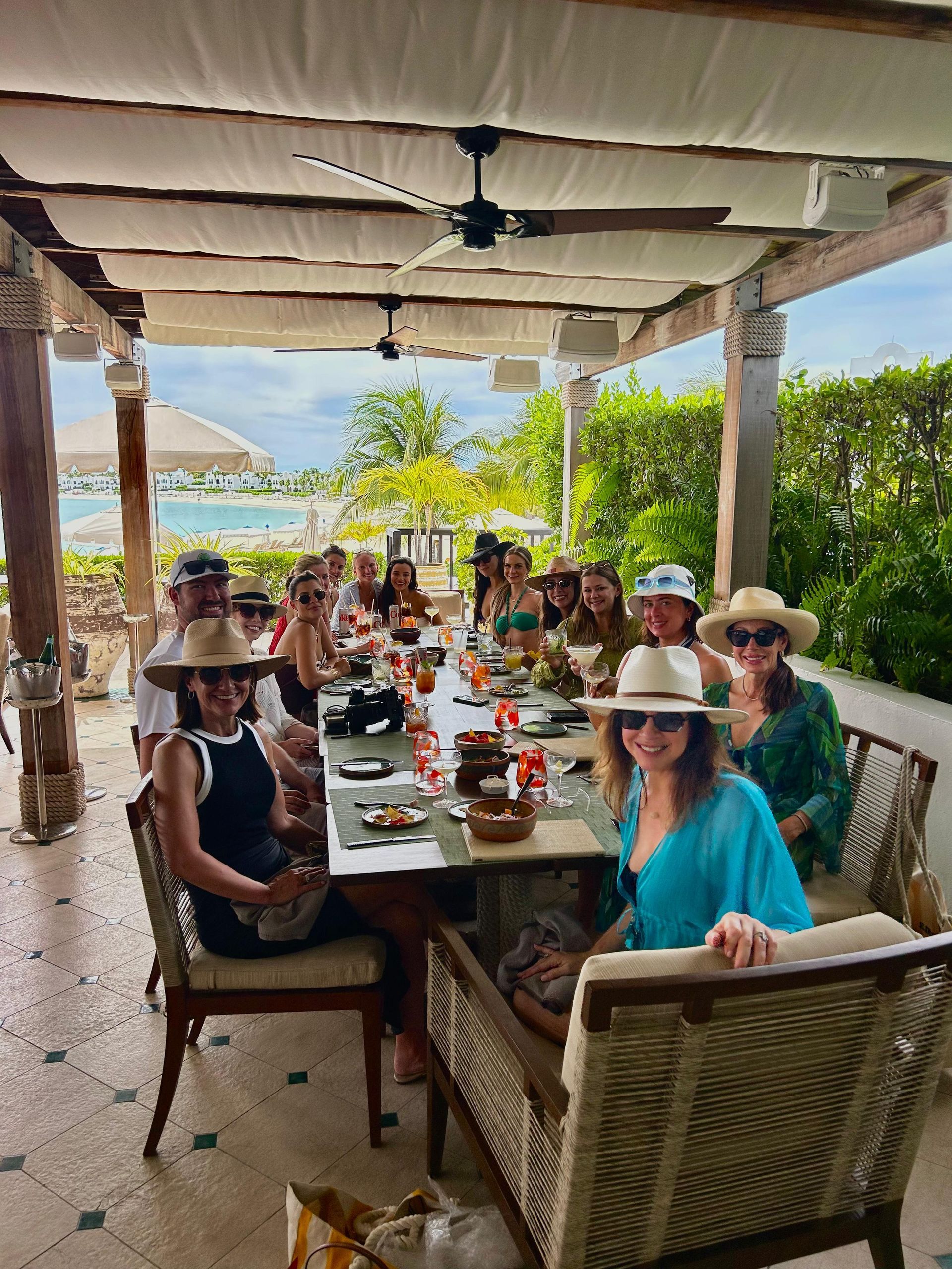 A group of people seated at a long table, eating outdoors under a shaded patio overlooking the ocean.