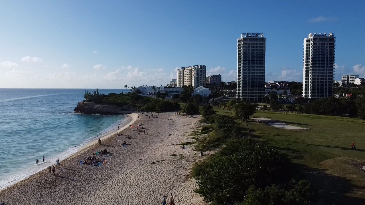 Beach scene with ocean, sand, buildings, blue sky, and some people.