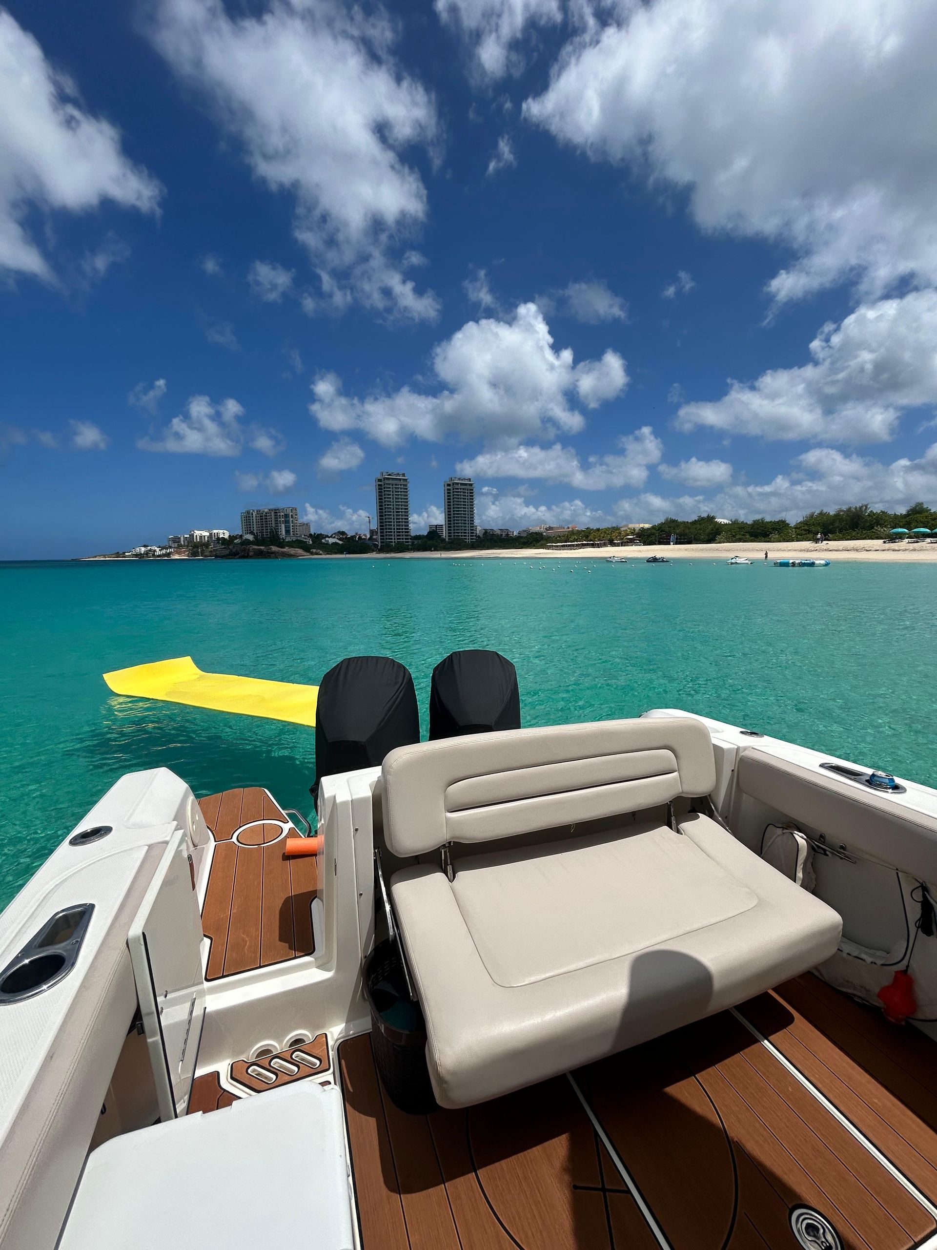 Boat on turquoise water, looking toward a beach and buildings under a blue sky.