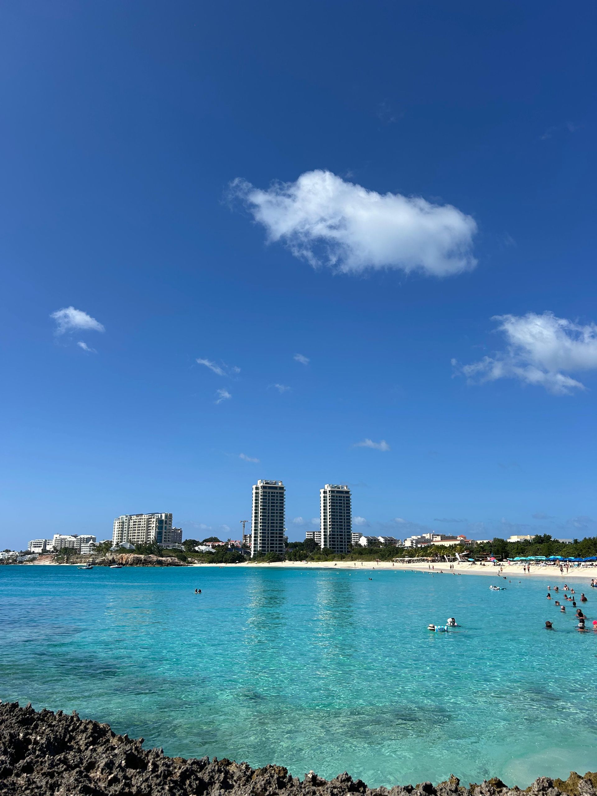 Turquoise water with beachgoers under a bright blue sky, with tall white buildings in the background.