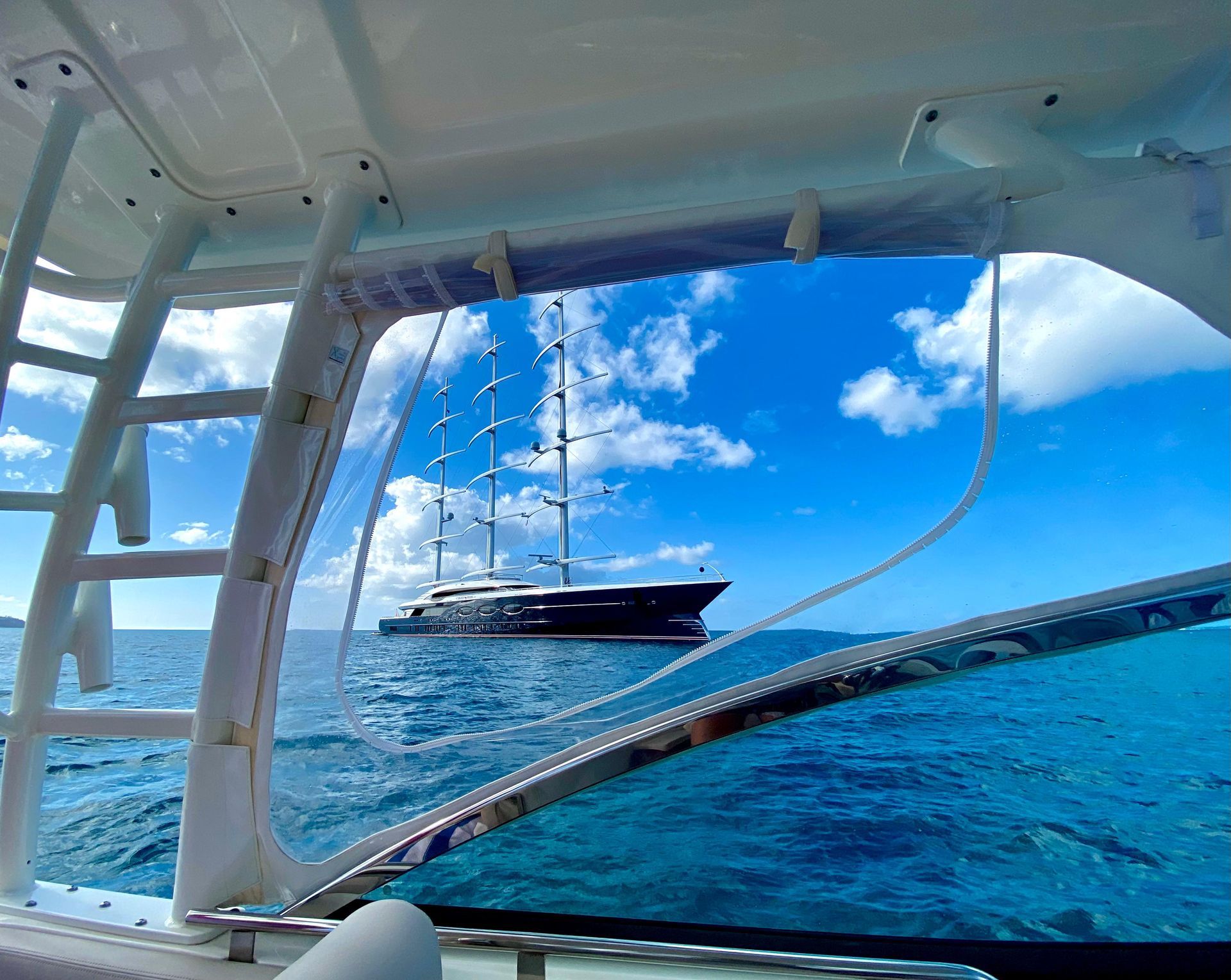 A large sailboat on the blue sea is seen through the window of a smaller boat, under a bright, sunny sky.