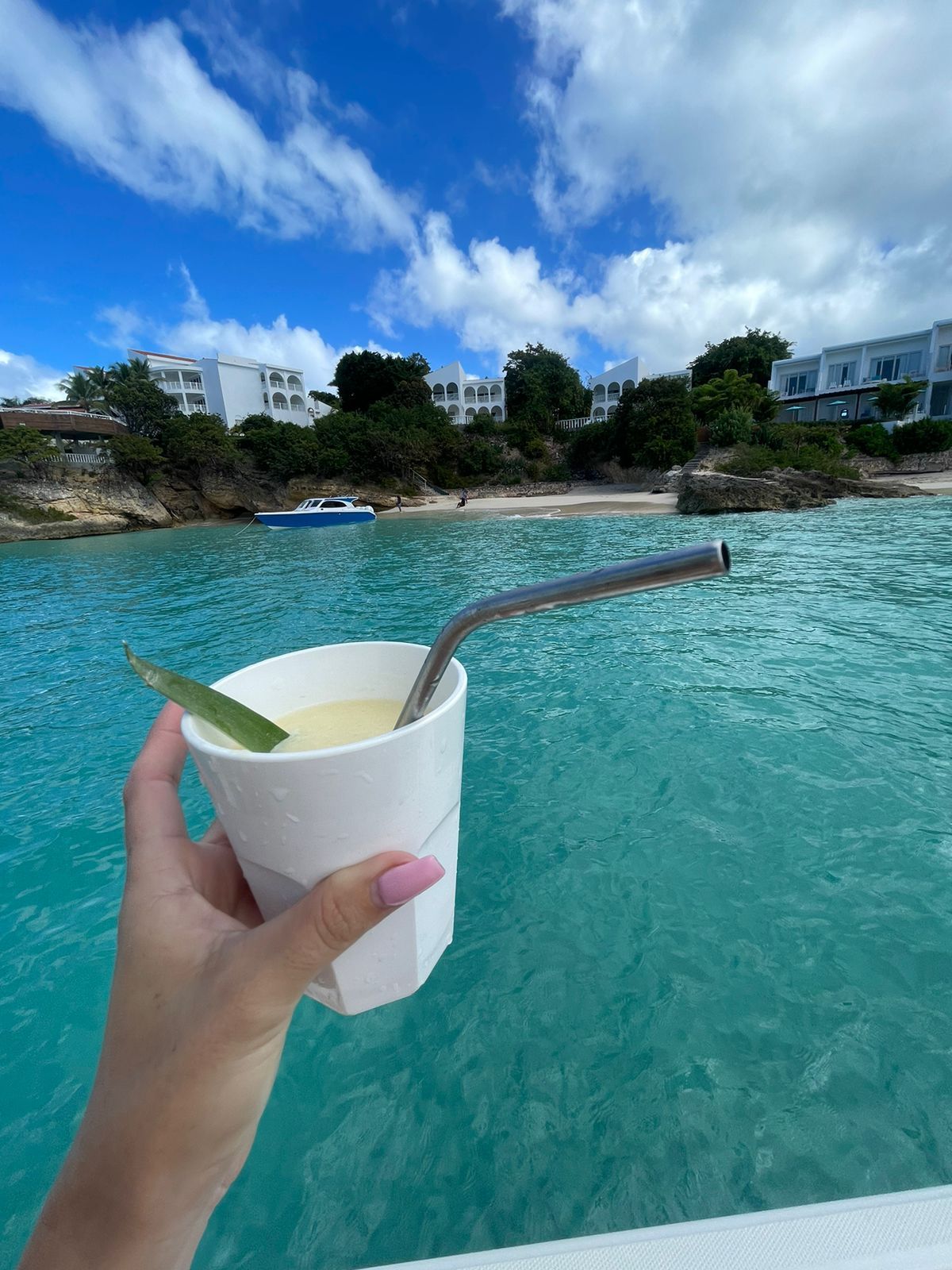 Hand holding drink with a metal straw, turquoise water, blue sky, and coastal buildings in the background.