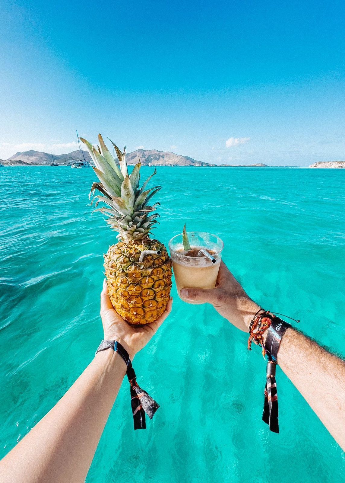 Hands holding a pineapple and cocktail in turquoise water, bright blue sky.