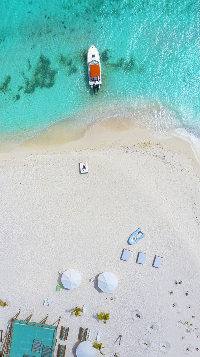Boat near a white sand beach with turquoise water. Lounge chairs, umbrellas, and small boats are also present.