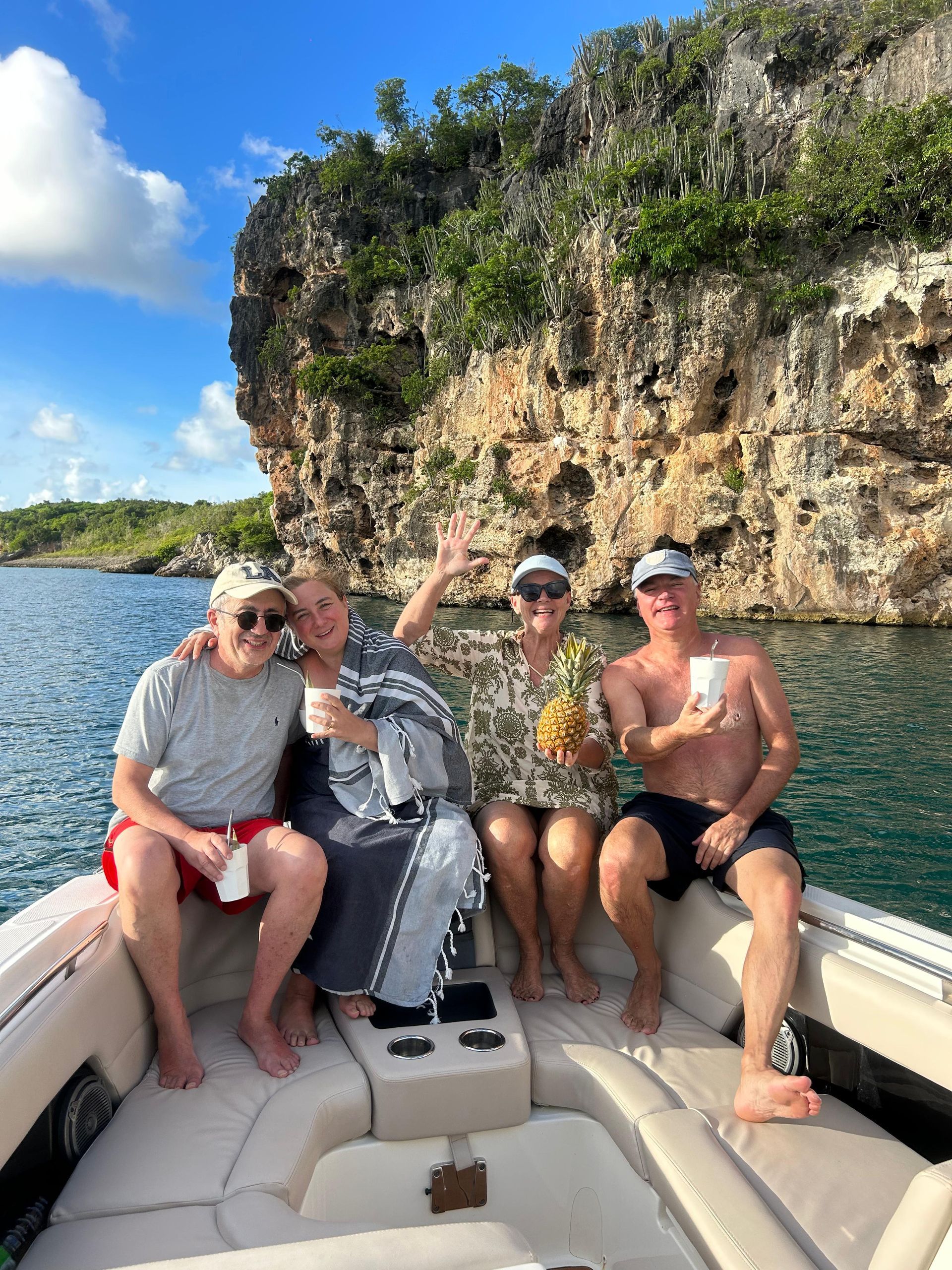 Four people wave from a boat on blue water, near a rocky cliff.