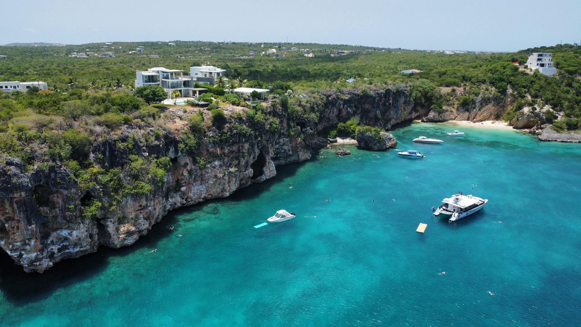 Coastal view: boats on turquoise water near a cliff with buildings, trees, and a beach.