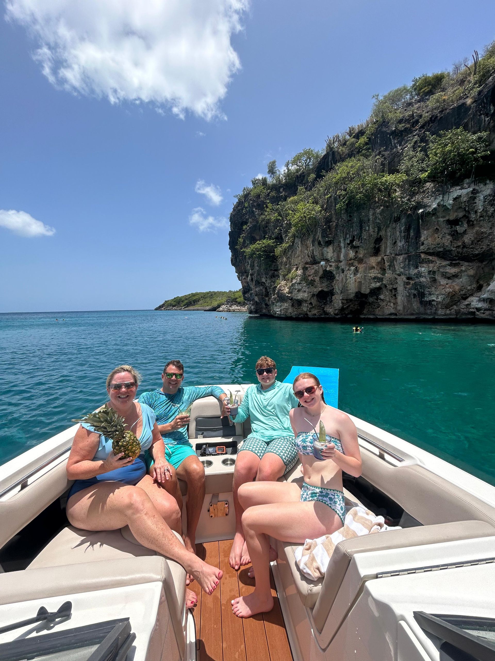 Four people on a boat with clear turquoise water near a rocky cliff on a sunny day.